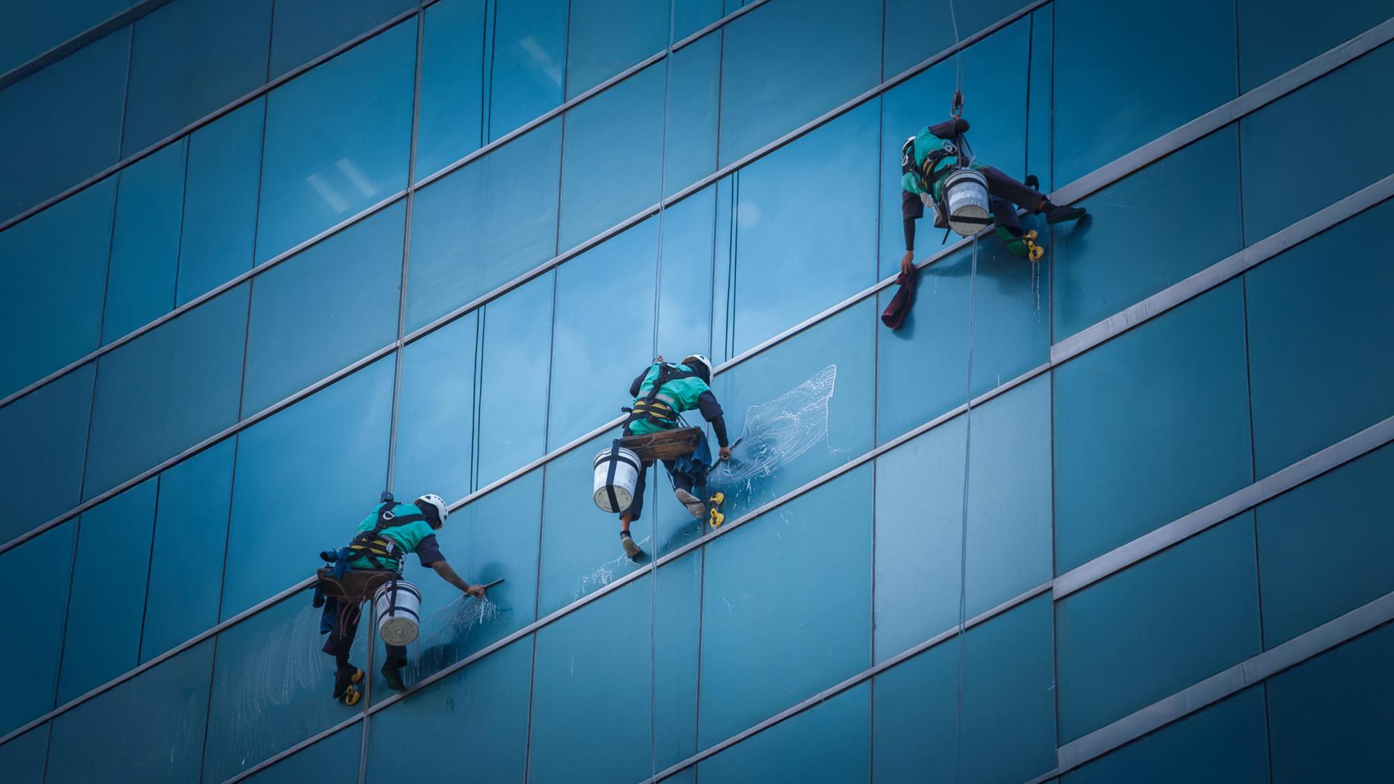 Three window washers in harnesses and helmets are cleaning the glass windows of a tall building, using ropes and equipment to access the high areas.